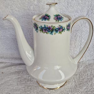 May include: White porcelain coffee pot with a floral design in shades of blue and purple. The pot has a gold trim around the lid and base.