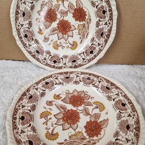 Könnte beinhalten: Zwei Vintage-Keramikteller mit cremefarbenem Hintergrund. Jeder Teller hat ein Blumendesign in Braun-, Orange- und Gelbtönen. Die Ränder haben einen dekorativen, erhabenen Rand.