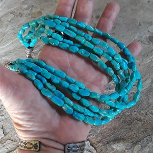 May include: A turquoise beaded necklace with multiple strands. The necklace features oval-shaped turquoise beads in varying shades of blue. The necklace is held in a hand, showcasing the vibrant color and texture of the stones. The necklace has silver-colored clasps.