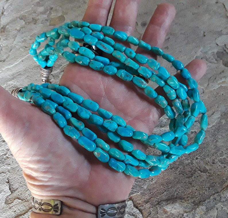 May include: A turquoise beaded necklace with multiple strands. The necklace features oval-shaped turquoise beads in varying shades of blue. The necklace is held in a hand, showcasing the vibrant color and texture of the stones. The necklace has silver-colored clasps.