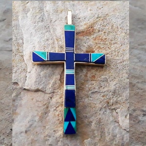 K&ouml;nnte beinhalten: Ein goldfarbener Kreuzanh&auml;nger mit eingelegten blauen und t&uuml;rkisfarbenen Steinen. Das Kreuz hat einen vertikalen Balken und horizontale Arme mit abwechselnden Abschnitten in Dunkelblau und T&uuml;rkis. Der Anh&auml;nger liegt auf einer strukturierten, hellfarbigen Steinoberfl&auml;che.