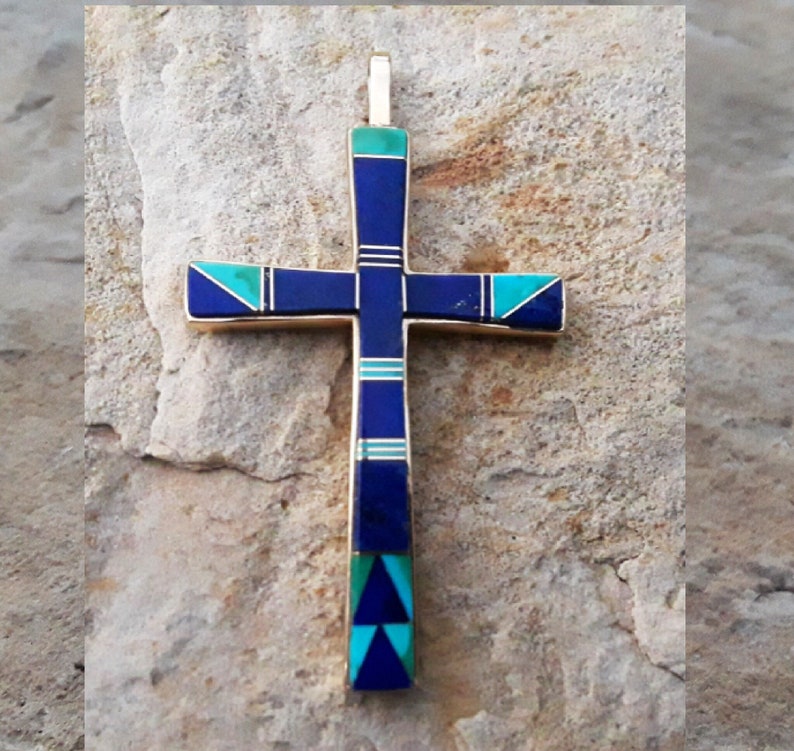 K&ouml;nnte beinhalten: Ein goldfarbener Kreuzanh&auml;nger mit eingelegten blauen und t&uuml;rkisfarbenen Steinen. Das Kreuz hat einen vertikalen Balken und horizontale Arme mit abwechselnden Abschnitten in Dunkelblau und T&uuml;rkis. Der Anh&auml;nger liegt auf einer strukturierten, hellfarbigen Steinoberfl&auml;che.