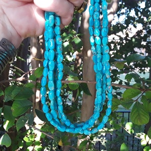May include: A multi-strand necklace featuring bright turquoise-colored beads. The beads are oval-shaped and strung together, creating a layered effect. The necklace is held up against a backdrop of green foliage.
