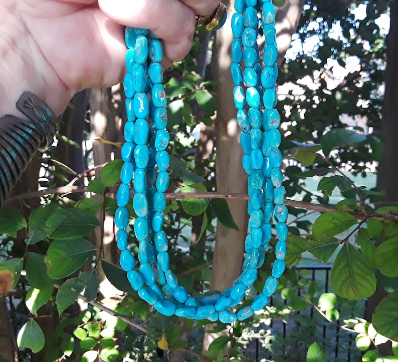 May include: A multi-strand necklace featuring bright turquoise-colored beads. The beads are oval-shaped and strung together, creating a layered effect. The necklace is held up against a backdrop of green foliage.