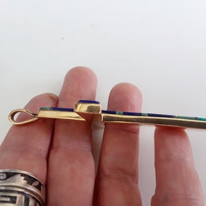 K&ouml;nnte beinhalten: Ein goldfarbener Kreuzanh&auml;nger mit eingelegten blauen und t&uuml;rkisfarbenen Steinen. Die Kreuzform wird von einer Hand gehalten, wodurch das detaillierte Design und die Handwerkskunst des Schmuckst&uuml;cks hervorgehoben werden.