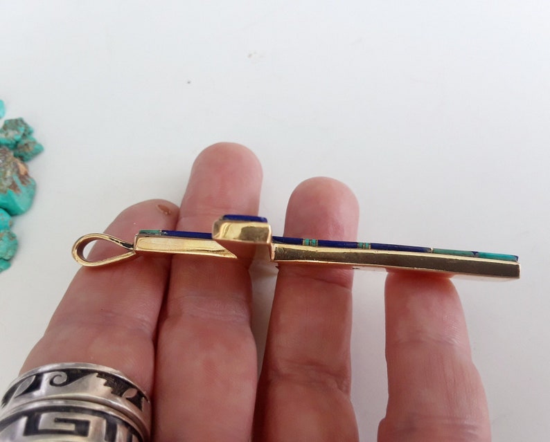 K&ouml;nnte beinhalten: Ein goldfarbener Kreuzanh&auml;nger mit eingelegten blauen und t&uuml;rkisfarbenen Steinen. Die Kreuzform wird von einer Hand gehalten, wodurch das detaillierte Design und die Handwerkskunst des Schmuckst&uuml;cks hervorgehoben werden.