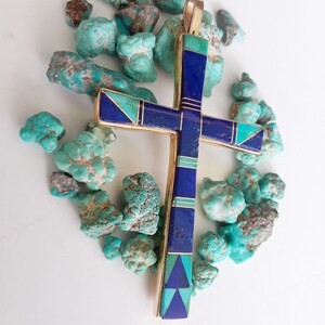 K&ouml;nnte beinhalten: Vergoldeter Kreuzanh&auml;nger mit Einlagen aus blauem Lapislazuli und T&uuml;rkissteinen. Das Kreuz ist von rohen T&uuml;rkissteinen umgeben. Der Anh&auml;nger hat eine goldene &Ouml;se f&uuml;r eine Halskette.