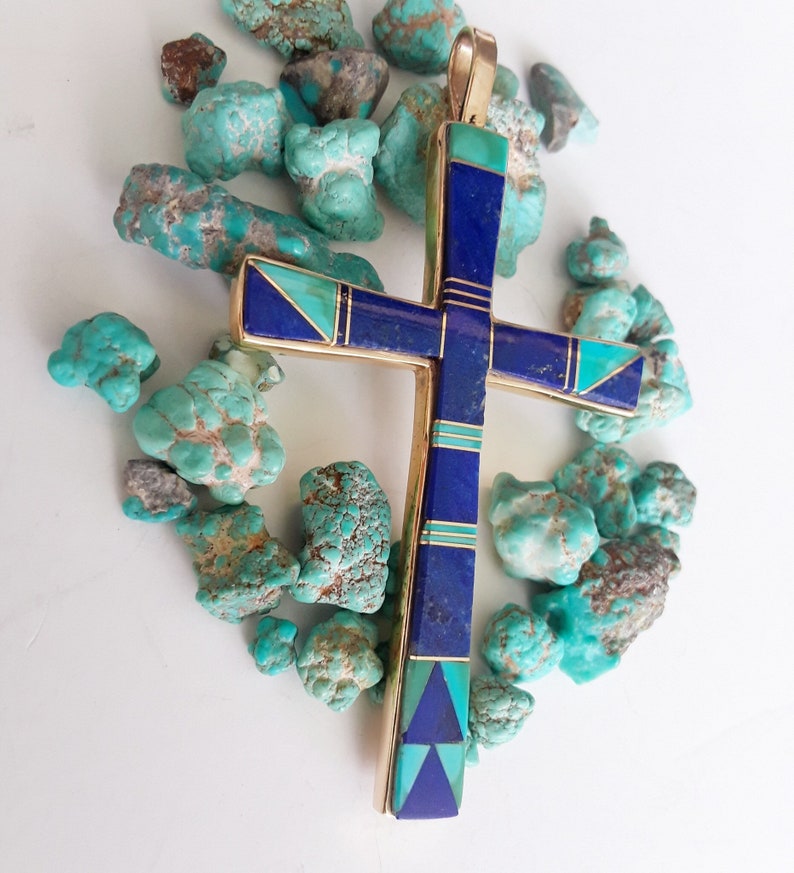 K&ouml;nnte beinhalten: Vergoldeter Kreuzanh&auml;nger mit Einlagen aus blauem Lapislazuli und T&uuml;rkissteinen. Das Kreuz ist von rohen T&uuml;rkissteinen umgeben. Der Anh&auml;nger hat eine goldene &Ouml;se f&uuml;r eine Halskette.