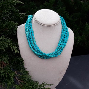 May include: A multi-strand turquoise necklace displayed on a white bust. The necklace features five strands of polished, irregular turquoise beads. The beads are a vibrant, consistent turquoise color. The background is a blurred mix of green foliage.