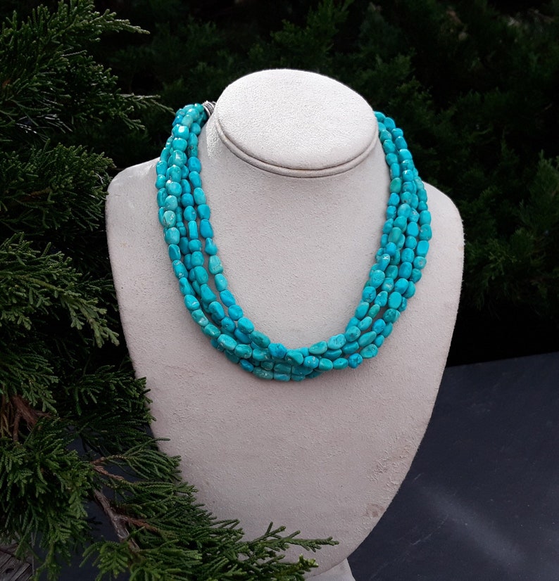 May include: A multi-strand turquoise necklace displayed on a white bust. The necklace features five strands of polished, irregular turquoise beads. The beads are a vibrant, consistent turquoise color. The background is a blurred mix of green foliage.