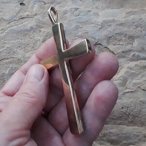 K&ouml;nnte beinhalten: Ein goldfarbener Kreuzanh&auml;nger mit einer Schlaufe oben f&uuml;r eine Kette. Das Kreuz hat ein schlichtes, klassisches Design mit poliertem Finish. Der Anh&auml;nger wird vor einem strukturierten, hellbraunen Hintergrund gehalten.