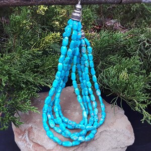 May include: A turquoise beaded necklace with multiple strands. The necklace features oval-shaped beads in a vibrant blue-green hue. The strands are connected at the top with silver-colored metal accents. The necklace is displayed on a rock with a backdrop of green foliage.