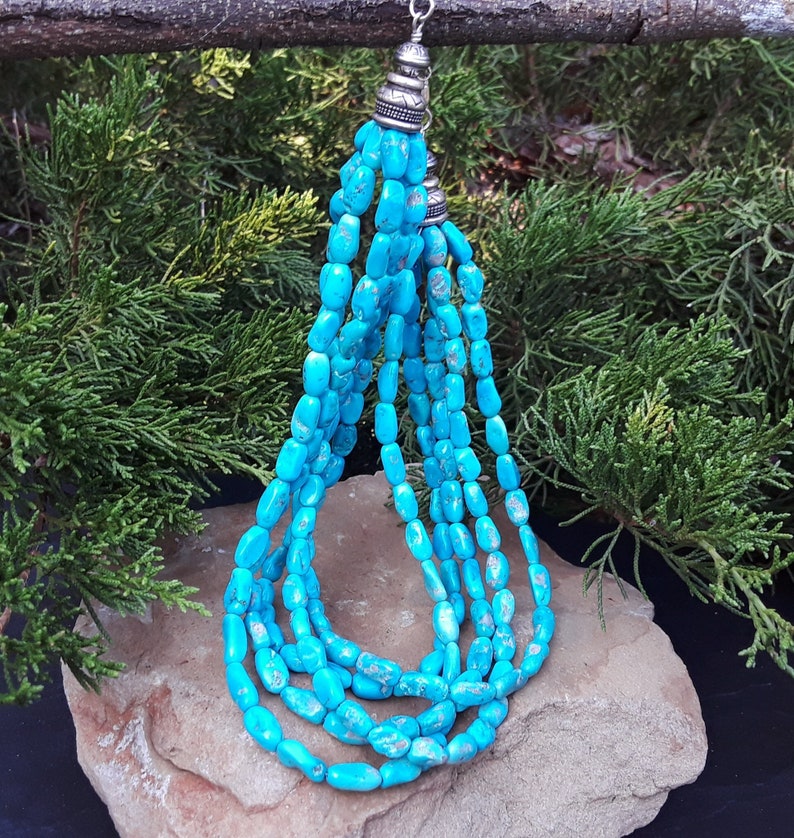 May include: A turquoise beaded necklace with multiple strands. The necklace features oval-shaped beads in a vibrant blue-green hue. The strands are connected at the top with silver-colored metal accents. The necklace is displayed on a rock with a backdrop of green foliage.