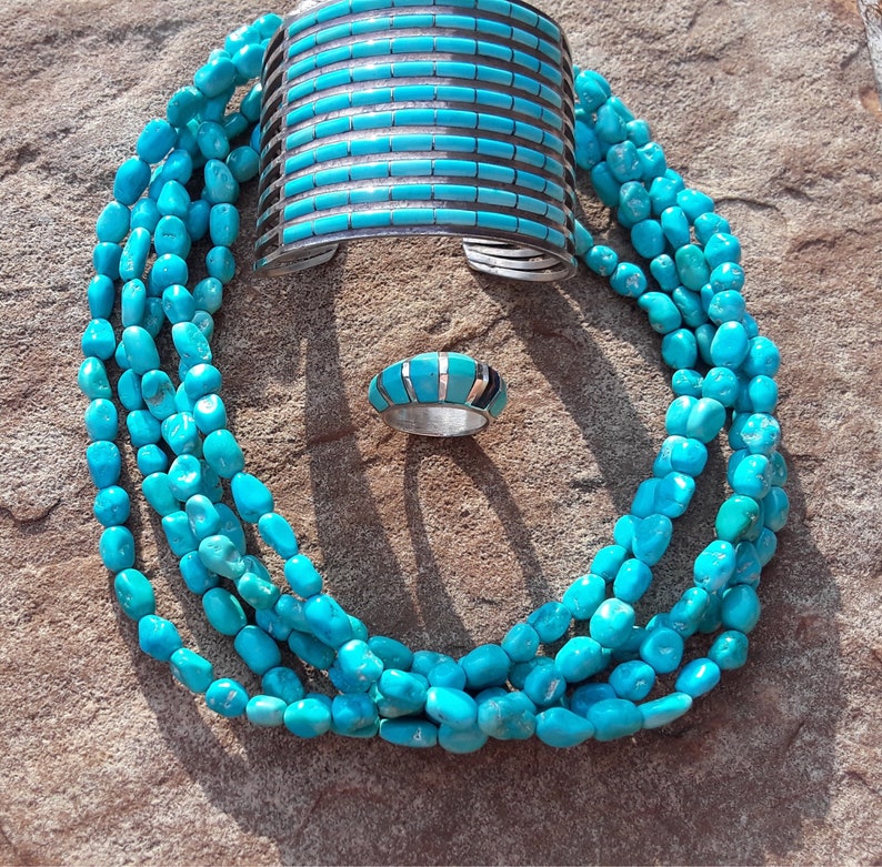 May include: A turquoise jewelry set featuring a multi-strand beaded necklace, a wide cuff bracelet, and a ring. The bracelet and ring have turquoise inlay designs. The jewelry is displayed on a textured, brown surface.