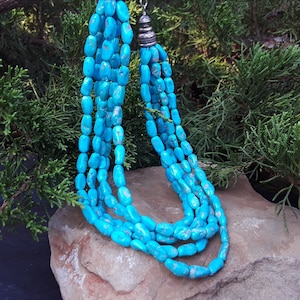 May include: A multi-strand turquoise necklace with a silver-toned clasp. The necklace features polished, oval-shaped turquoise beads in varying shades of blue. The necklace is displayed on a stone surface with a backdrop of green foliage.