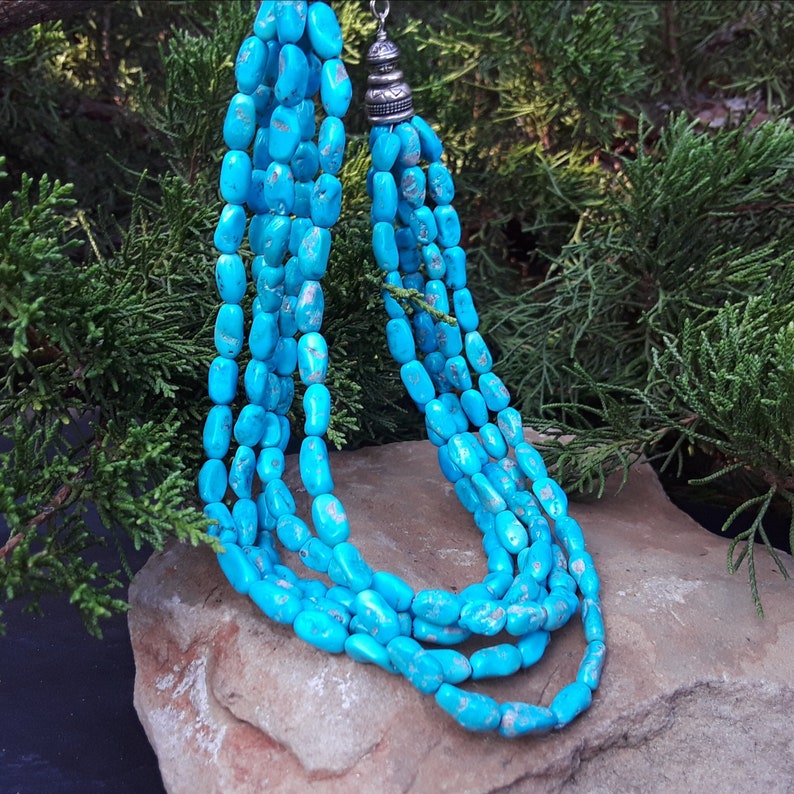May include: A multi-strand turquoise necklace with a silver-toned clasp. The necklace features polished, oval-shaped turquoise beads in varying shades of blue. The necklace is displayed on a stone surface with a backdrop of green foliage.