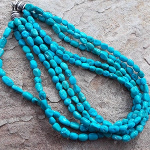 May include: A turquoise necklace with multiple strands of polished, oval-shaped beads. The necklace has a silver clasp and is displayed on a textured, neutral-toned surface. The beads are a vibrant, eye-catching blue.
