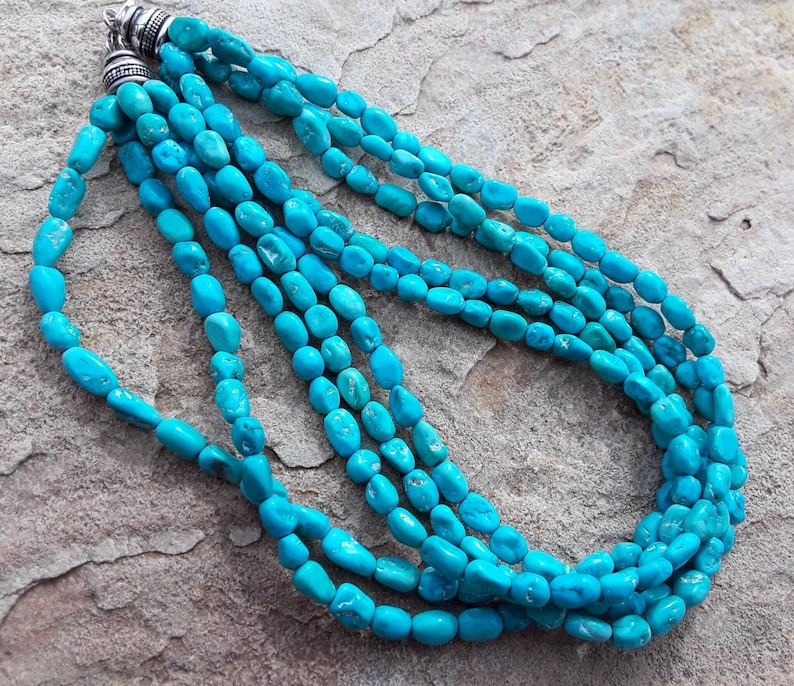 May include: A turquoise necklace with multiple strands of polished, oval-shaped beads. The necklace has a silver clasp and is displayed on a textured, neutral-toned surface. The beads are a vibrant, eye-catching blue.