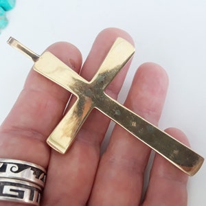 K&ouml;nnte beinhalten: Ein gro&szlig;er, goldfarbener Kreuzanh&auml;nger mit rechteckigem Design. Das Kreuz hat oben eine Schlaufe f&uuml;r eine Kette. Der Anh&auml;nger wird in einer Hand gehalten, mit einer t&uuml;rkisfarbenen Perlenkette und Silberringen im Hintergrund.
