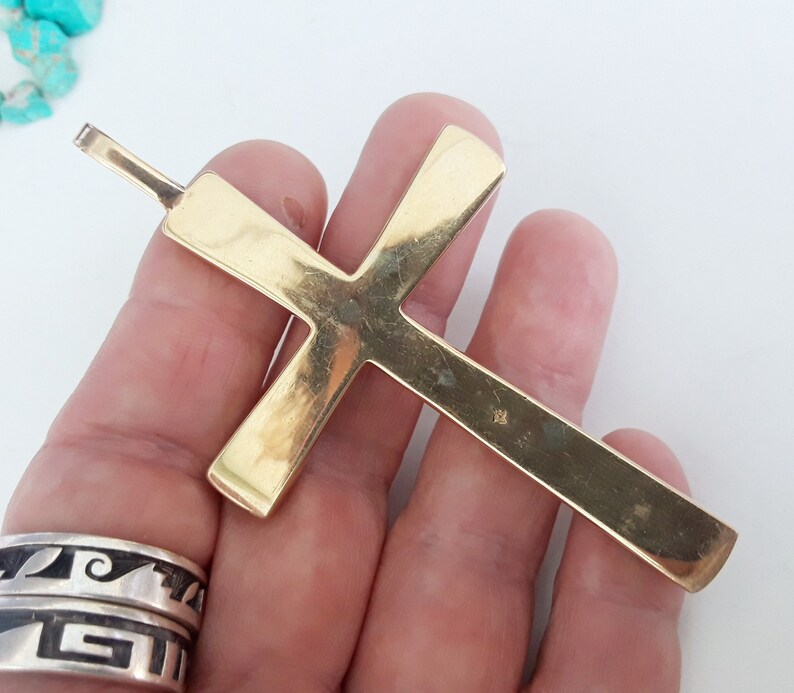 K&ouml;nnte beinhalten: Ein gro&szlig;er, goldfarbener Kreuzanh&auml;nger mit rechteckigem Design. Das Kreuz hat oben eine Schlaufe f&uuml;r eine Kette. Der Anh&auml;nger wird in einer Hand gehalten, mit einer t&uuml;rkisfarbenen Perlenkette und Silberringen im Hintergrund.