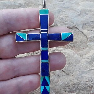 K&ouml;nnte beinhalten: Ein silberfarbener Kreuzanh&auml;nger mit eingelegten blauen und t&uuml;rkisfarbenen Steinen. Das Kreuz hat ein rechteckiges Design mit geometrischen Mustern und eine Schlaufe oben f&uuml;r eine Kette. Die Steine sind symmetrisch angeordnet.