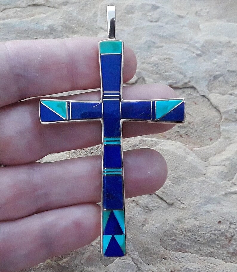 K&ouml;nnte beinhalten: Ein silberfarbener Kreuzanh&auml;nger mit eingelegten blauen und t&uuml;rkisfarbenen Steinen. Das Kreuz hat ein rechteckiges Design mit geometrischen Mustern und eine Schlaufe oben f&uuml;r eine Kette. Die Steine sind symmetrisch angeordnet.