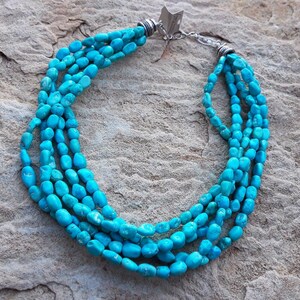 May include: A turquoise beaded necklace with multiple strands. The necklace features bright blue-green stones and a silver-toned clasp. The beads are irregularly shaped and arranged in a cascading design, creating a layered effect.