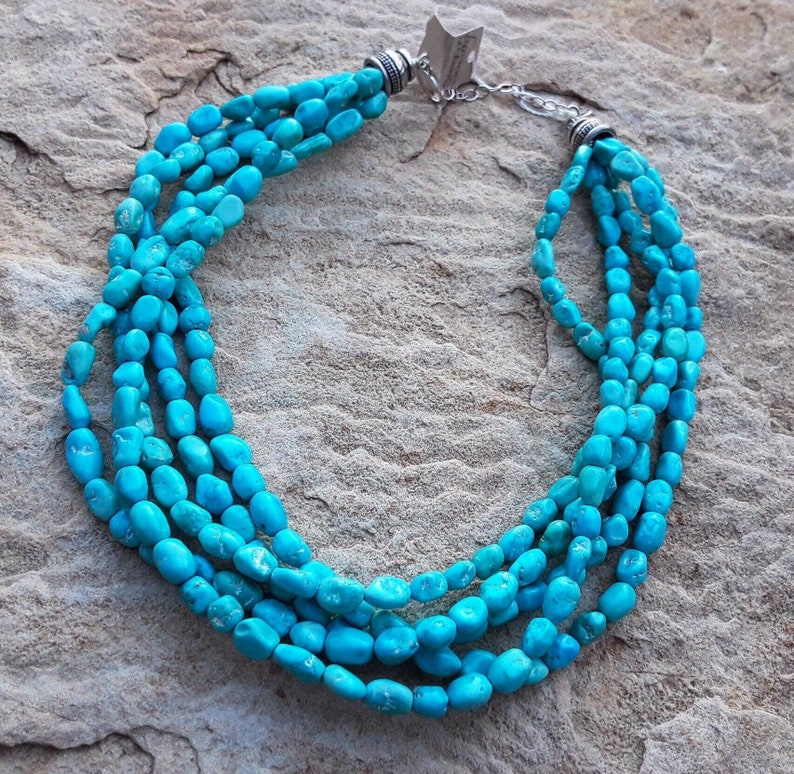 May include: A turquoise beaded necklace with multiple strands. The necklace features bright blue-green stones and a silver-toned clasp. The beads are irregularly shaped and arranged in a cascading design, creating a layered effect.