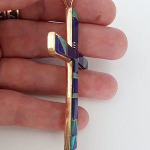 K&ouml;nnte beinhalten: Goldener Anh&auml;nger in Form eines Kreuzes. Das Kreuz hat eingelegte Abschnitte aus blauen, gr&uuml;nen und violetten Steinen. Der Anh&auml;nger hat oben eine Schlaufe f&uuml;r eine Kette.
