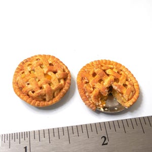 May include: Two miniature fruit pies with golden-brown lattice crusts, one whole and one with a slice removed, positioned above a metal ruler showing a diameter of approximately 1 inch for each pie, set against a plain white background.