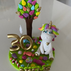Peut inclure: Une décoration de gâteau représentant une licorne blanche avec une crinière arc-en-ciel et une corne dorée, un arbre brun aux feuilles colorées, un chiffre 30 doré et des bonbons sur une base verte. Le nom AMELIE est écrit en lettres dorées.