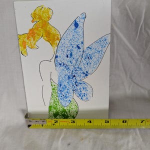 May include: A white card featuring a whimsical illustration of a fairy. The fairy has yellow hair, blue and green wings, and a black outline. A yellow measuring tape is placed at the bottom of the card, showing measurements in inches.
