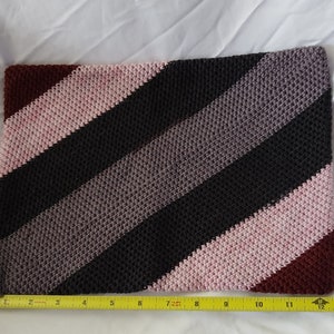 May include: A crocheted striped pillow with diagonal stripes in shades of burgundy, pink, gray, and black. The pillow is approximately 12 inches long.