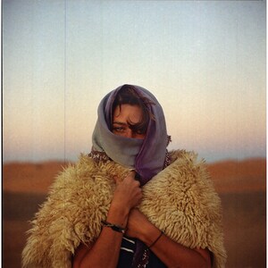 May include: A person wearing a purple and blue headscarf and a brown fur coat. The person is standing in a desert landscape.