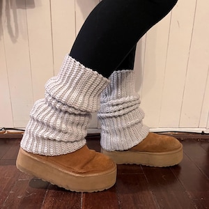 May include: A pair of grey crocheted leg warmers with a ribbed texture. The leg warmers are worn over brown suede boots with a thick platform sole.