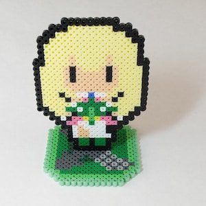 May include: A pixel art figurine of a cartoon character with blonde hair, a green dress, and a pink bow. The figurine is standing on a green base with a black border.