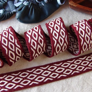 Peut inclure: Gros plan sur un ruban tissé bordeaux et blanc avec un motif de losanges répétés. Le ruban est enroulé et posé sur une surface neutre. En arrière-plan, une paire de chaussures en cuir noir.