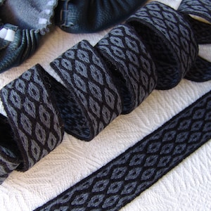 May include: A rolled length of black and gray patterned ribbon with a diamond design. The ribbon is made of a woven material and is displayed on a white textured surface. A pair of dark blue leather baby shoes are in the background.