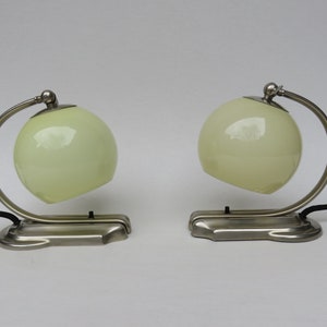 May include: Two matching Art Deco-style table lamps. Each lamp features a spherical, pale green glass shade supported by a curved, brushed metal arm. The base is a rectangular shape with a brushed metal finish. Black cords extend from the base.