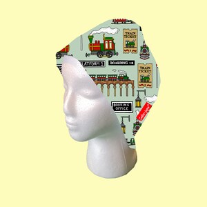 May include: A green and white fabric headband with a repeating pattern of cartoon trains, train tickets, and clouds. The text "Train Ticket" and "Booking Office" are visible on the headband.