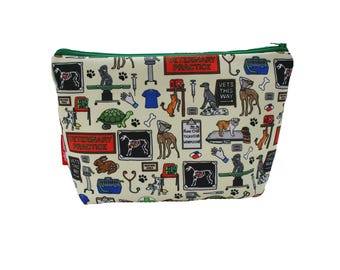 May include: A cream-colored zippered cosmetic bag with a green zipper and trim. The bag is covered in a repeating pattern of veterinary-themed illustrations, including dogs, cats, stethoscopes, and the words "Veterinary Practice."