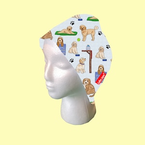 May include: A white surgical cap with a blue and white pattern of doodle dogs. The dogs are wearing collars and some are holding dog toys. The text "Sailor Love" is printed in red on the cap.