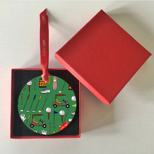May include: A round green bauble with a golf theme. It features golf carts, golf clubs, golf balls, and other golf-related items. The bauble is in a red box with a black interior.