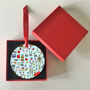 以下が含まれることがあります： 様々な医療記号と白いコートが描かれた、丸い青と白のクリスマスオーナメント。オーナメントは、黒い内側の赤い箱に入っています。オーナメントには、「Selina Jayne」という文字が印刷された赤いリボンが付いています。