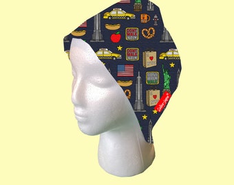 May include: A blue and yellow patterned fabric headband with a New York City theme. The headband features images of taxis, hot dogs, the Statue of Liberty, and the Empire State Building. The text "I Love NYC" is also included on the headband.