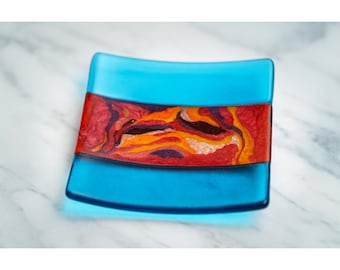 May include: A square, turquoise glass dish with a vibrant, abstract design. The central band features a mix of red, orange, and yellow hues, creating a striking contrast against the blue. The dish is a decorative item.