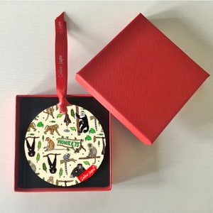 May include: A round Christmas ornament with a monkey pattern, presented in a red gift box. The ornament features various monkey illustrations and the word "MONKEYS". A red ribbon with the text "Selina-Jayne" is attached.