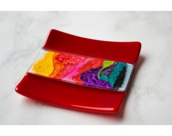 May include: A red, square-shaped dish with a rectangular, colorful insert. The insert features a vibrant mix of yellow, orange, pink, purple, green, and blue hues. The dish is likely made of glass or a similar material.