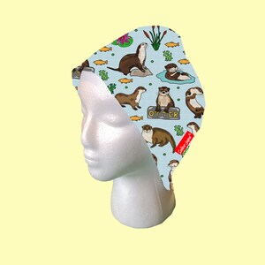 May include: A blue and white patterned fabric headband with a repeating design of otters, fish, and lily pads. The headband has the word "Otter" printed on it.