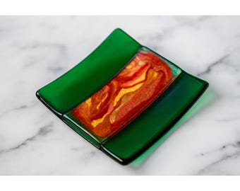 May include: A square, green glass dish with a central rectangular design in shades of red, orange, and gold. The dish has a slightly raised edge and sits on a white marble surface. This decorative dish is suitable for holding small items.